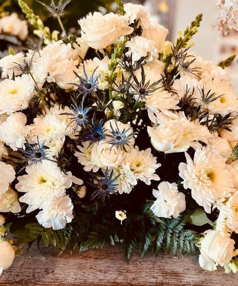 A floral arrangement featuring white roses, chrysanthemums, and greenery, accented with blue thistles.