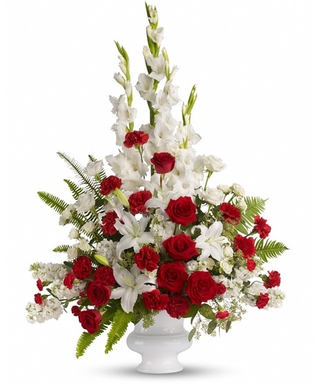 A tall floral arrangement with red roses, white lilies, white gladiolus, red carnations, and green ferns in a white vase.